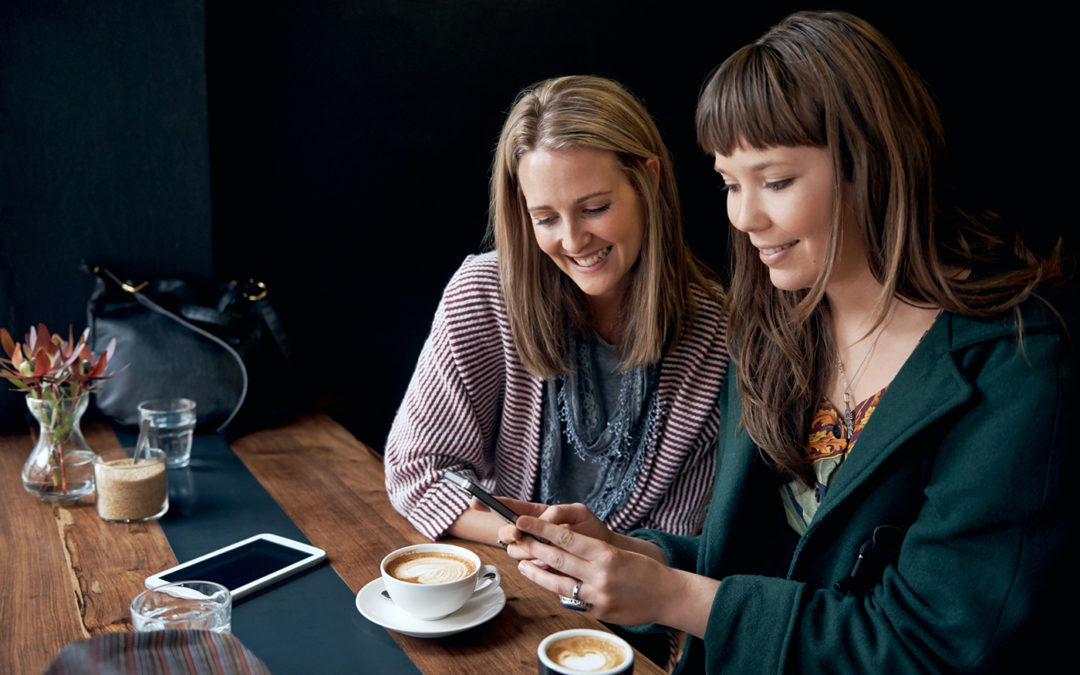 Two women smiling and looking at a smartphone at a café table with coffee cups.