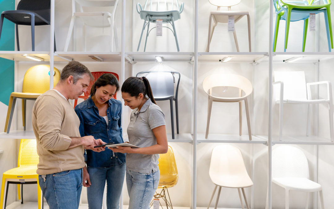 Three people looking at a tablet in a modern furniture store with chairs on display.
